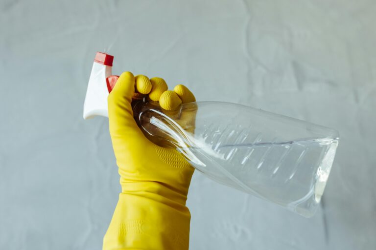 Close-up of a hand wearing yellow gloves holding a transparent spray bottle, isolated on a plain background.